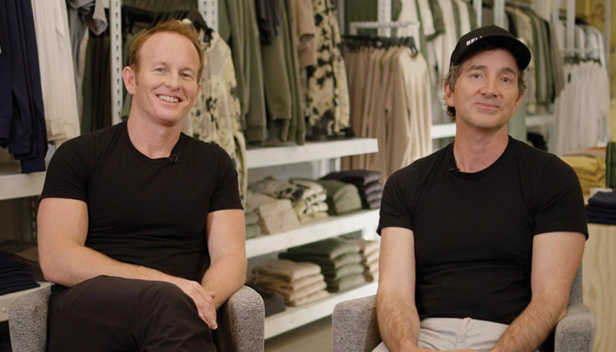 two men seated next to each other, apparel in background